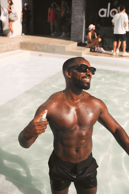 Man wearing Alo shorts standing knee-deep in a swimming pool, spraying his chest with Magnesium Reset Spray